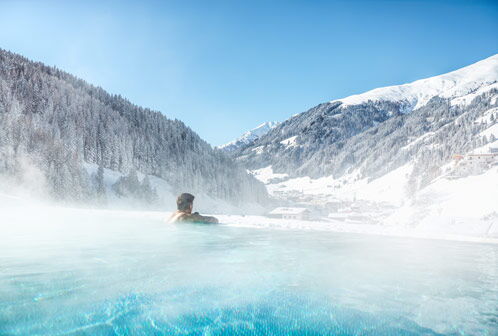 Mann in einem dampfendem Pool mit Blick auf verschneite Berge